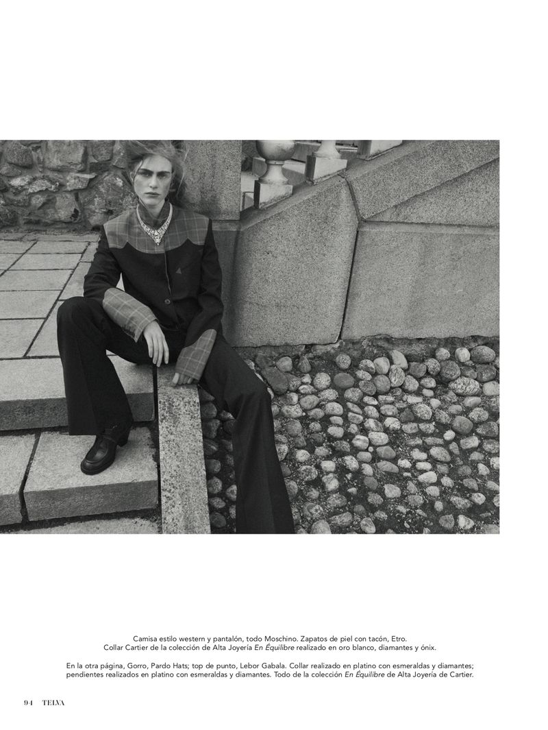 Monochrome portrait of a young adult man in a western-style suit with statement jewelry, sitting on a concrete stairway with a pebble ground.