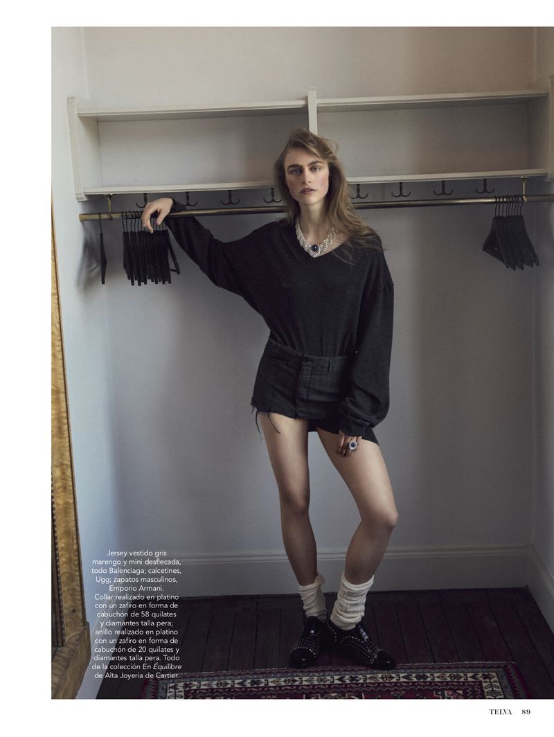 Woman poses in a dark V-neck sweater and denim shorts with white socks and black shoes in an indoor photo studio setting.