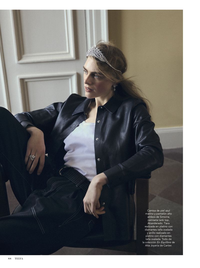 Woman with tiara sits in chair, wearing black leather jacket and dark jeans, illuminated by indoor lighting, showcasing a fashion editorial.