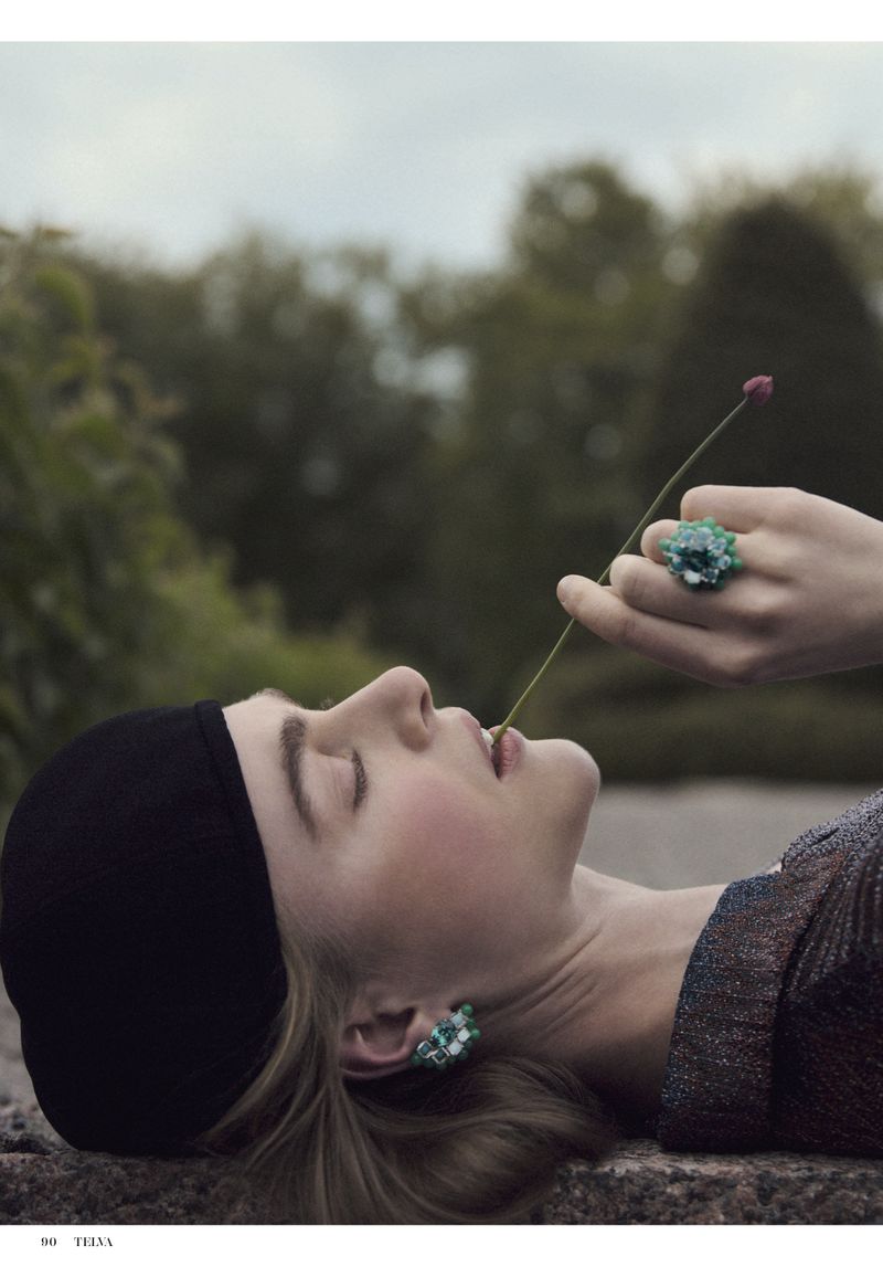 A woman lies with closed eyes, turquoise jewelry, and a flower stem held in her mouth, against a blurred background of trees.