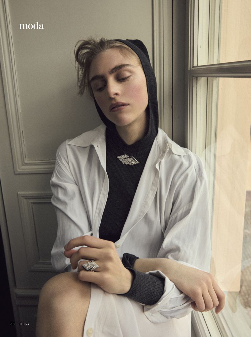 Fashion shot of a young woman in a white shirt and gray turtleneck with jewelry by a window in soft, diffused natural light.