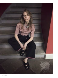 Model wearing a pink t-shirt and black trousers sitting on grey stone stairs framed by deep red walls.
