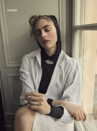 Model Hedvig Palm rests by a window wearing an open white button-down shirt over a black knit hood with diamond jewelry.
