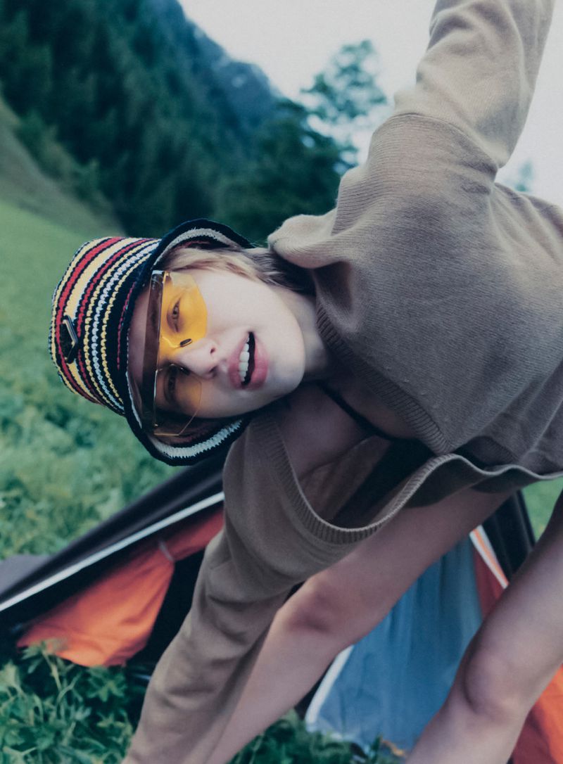 Young woman in a hat and glasses bending over in a grassy field with a mountain in the background and a tent in the foreground.