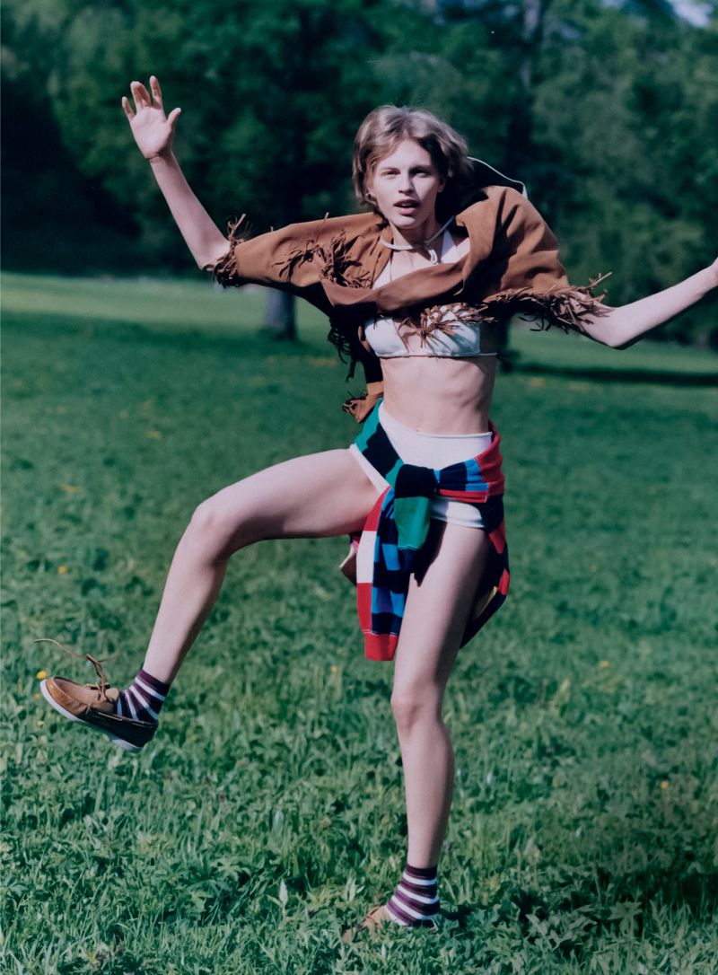 Young woman in vintage fashion poses outdoors in a green field wearing a fringed brown shawl, bra top, and striped socks with brown shoes.