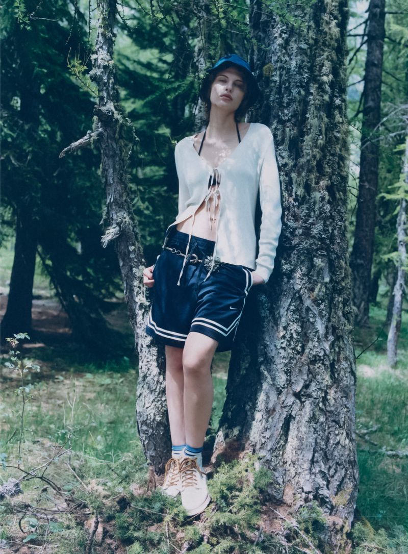 Adult female model poses in a forest wearing a sweater, shorts, bucket hat and sneakers leaning against a tree on a sunny day.