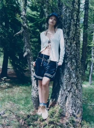 Adult female model poses in a forest wearing a sweater, shorts, bucket hat and sneakers leaning against a tree on a sunny day.