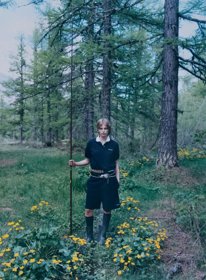 A person in outdoor fashion stands in a forest wearing a black polo shirt, black shorts, and boots, holding a fishing rod, nature setting.