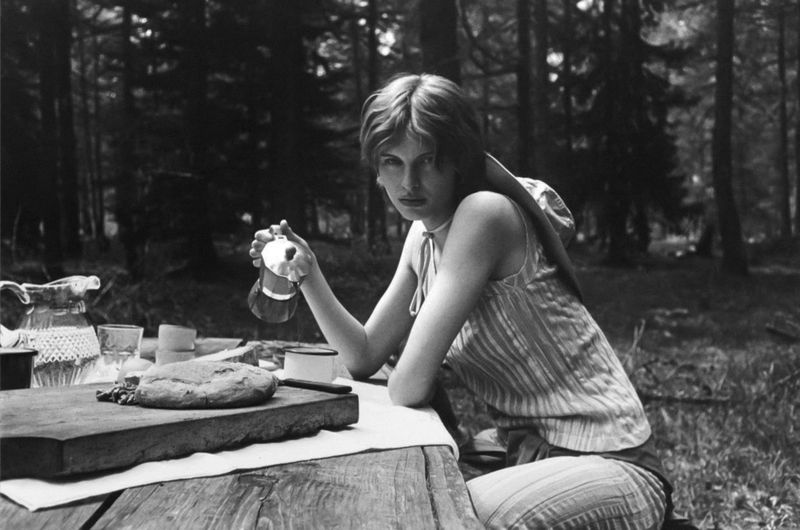 Adult female with short hair sits at a picnic table in a forest, holding a glass pitcher with bread, knife, glasses and trees.