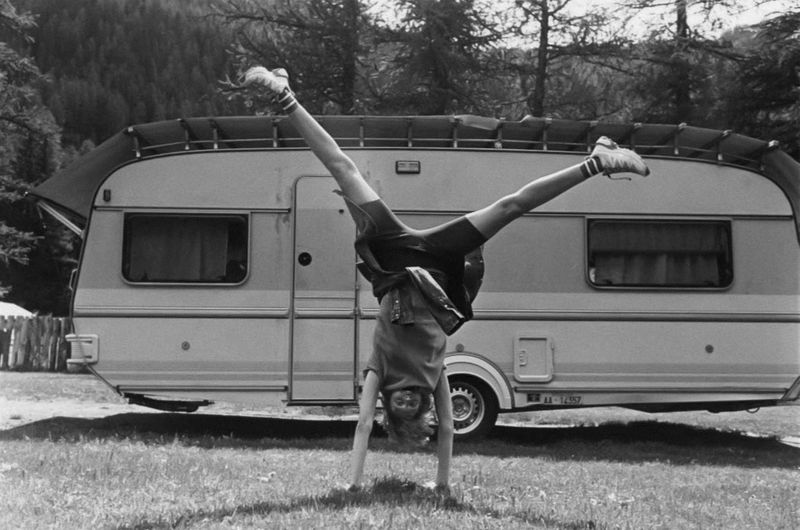 Child performs a handstand on the grass in front of a trailer on a sunny day. The background features green grass, trees, and sky.
