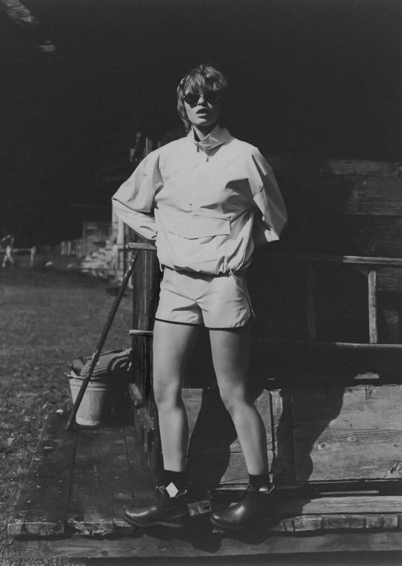 Monochrome shot of an adult standing on a wooden platform, wearing a jacket, shorts, sunglasses, and wooden shoes in an outdoor setting.