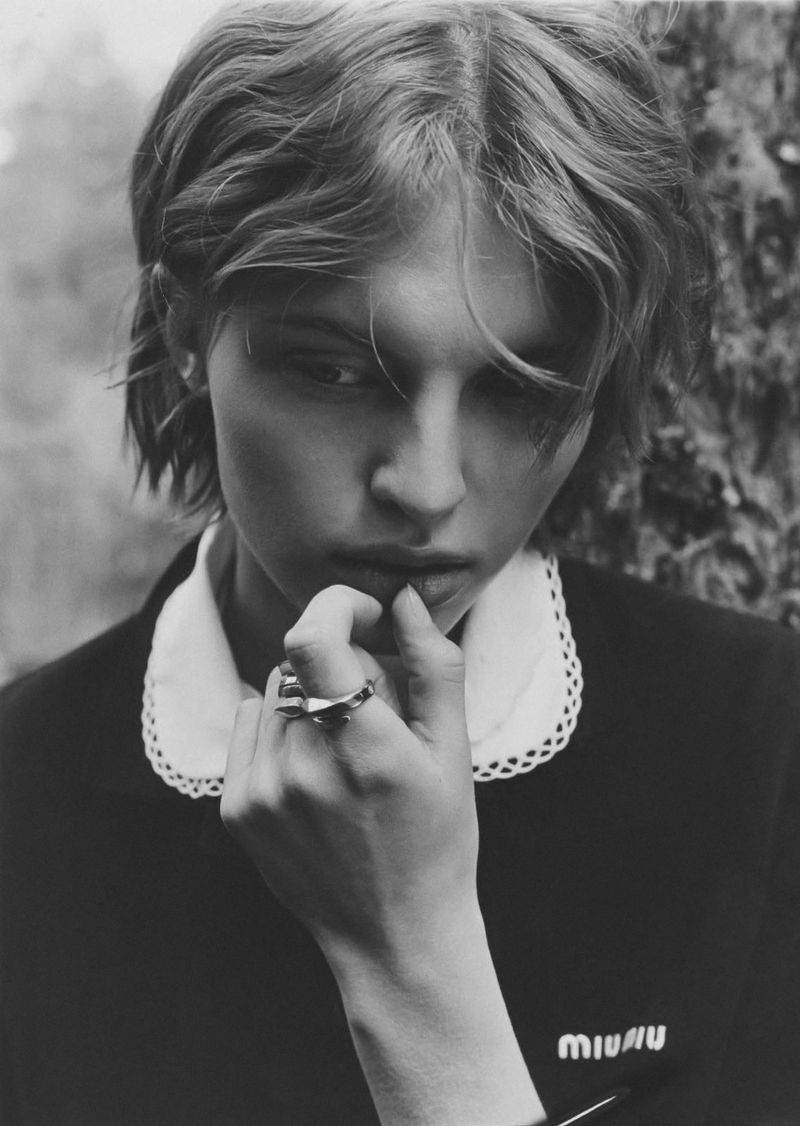 Monochrome portrait of a young adult woman with short wavy hair, wearing a collared shirt, in an outdoor woodland setting.