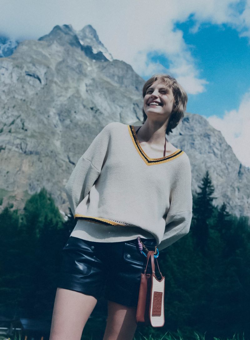 Model smiling in a cream v-neck knit sweater and black leather shorts against a dramatic alpine mountain backdrop.