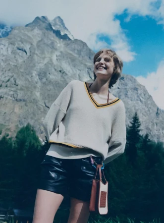 Model smiling in a cream v-neck knit sweater and black leather shorts against a dramatic alpine mountain backdrop.