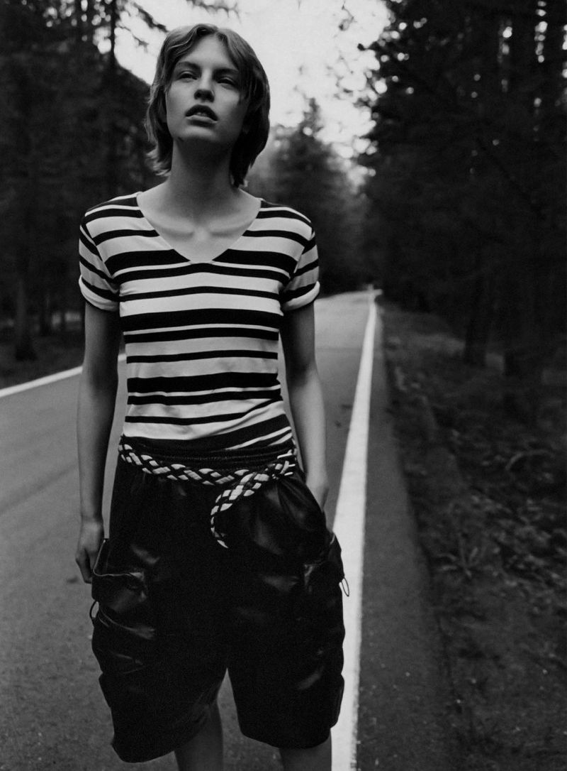 Woman stands on road in black and white photo. She wears a striped shirt, braided belt, and shorts, with trees lining the road.