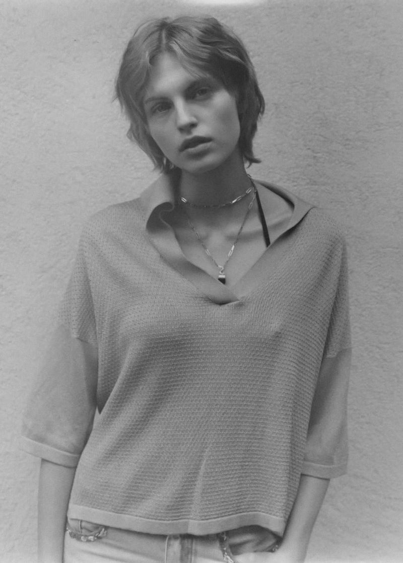 Monochrome portrait of a woman with short hair wearing a collared shirt and a layered necklace against a textured wall backdrop.