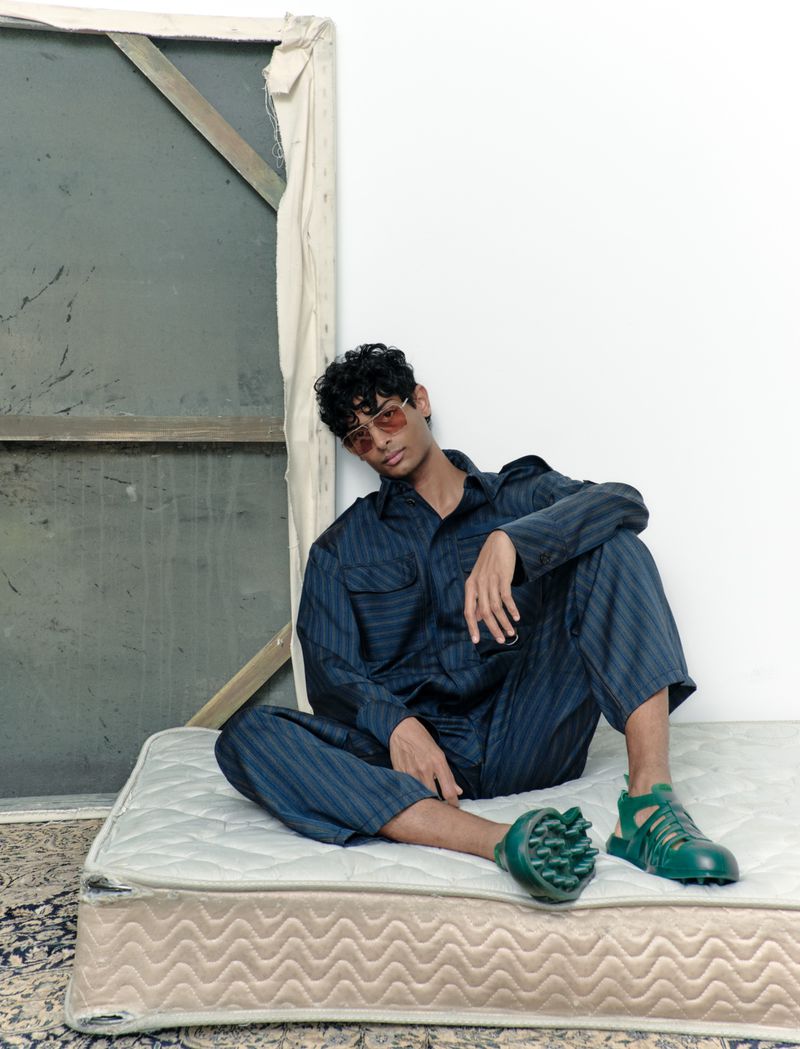 A young man with curly hair and sunglasses poses in a navy striped outfit and green shoes while sitting on an old mattress.
