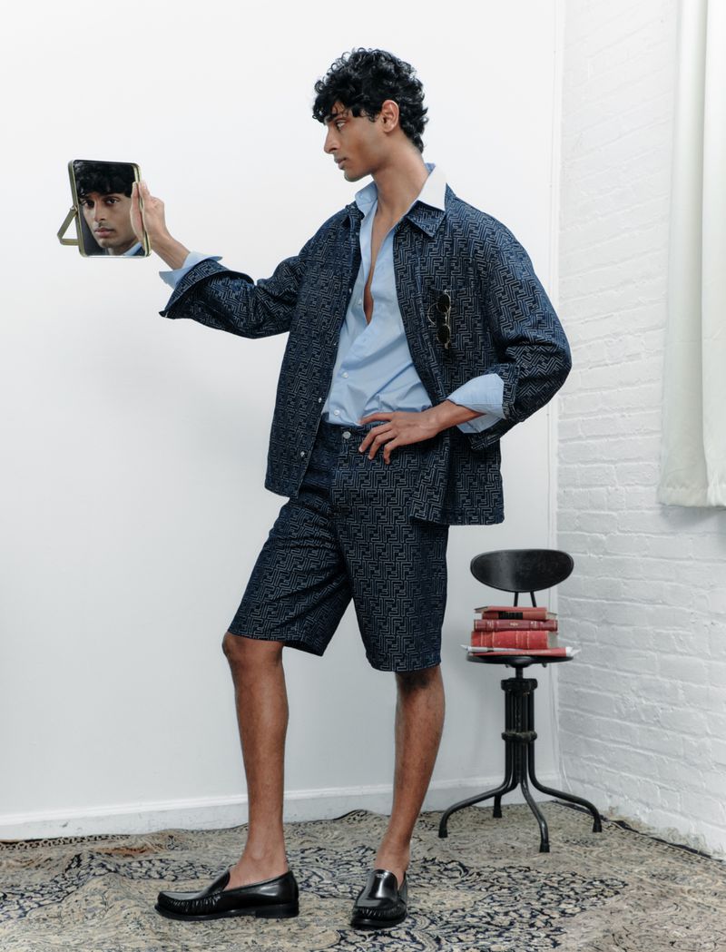 A South Asian adult male models a patterned blue suit with an open dress shirt, in an indoor setting, holding a mirror.