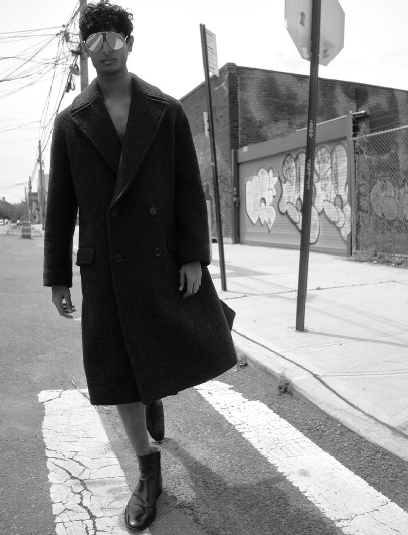 Model Pratik Shetty walks across an urban crosswalk wearing a long tailored dark coat and oversized sunglasses in black and white.