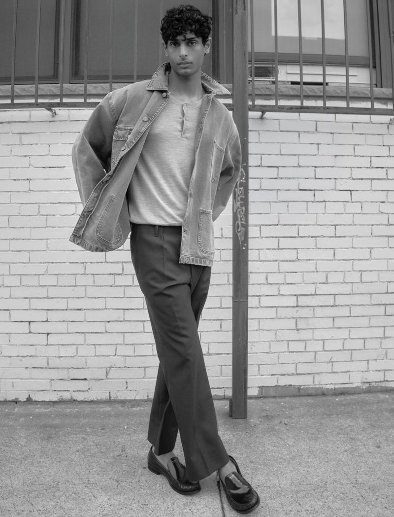 Model Pratik Shetty standing by a brick wall wearing an open jacket, henley shirt, trousers, and loafers in black and white.