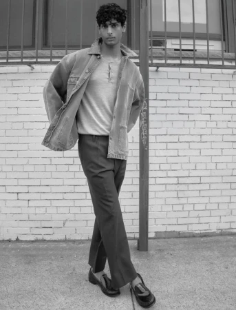 Model Pratik Shetty standing by a brick wall wearing an open jacket, henley shirt, trousers, and loafers in black and white.