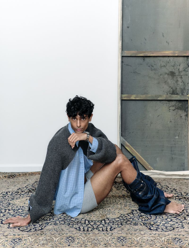 Man in unbuttoned shirt and jeans around lower legs sits on patterned rug in studio with gray panel background, facing camera.