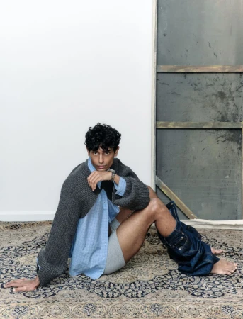 Man in unbuttoned shirt and jeans around lower legs sits on patterned rug in studio with gray panel background, facing camera.