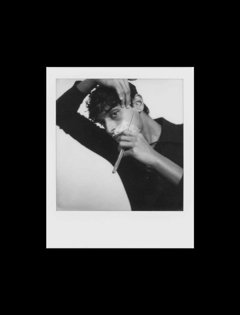 Black and white portrait of a young adult male shaving his face with a straight razor, depicted within a polaroid frame.