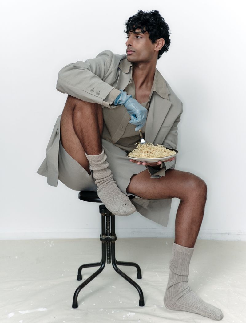 Young man in a tan jacket and shorts sitting on a stool, wearing socks and blue gloves, holding a plate of spaghetti in a white studio.