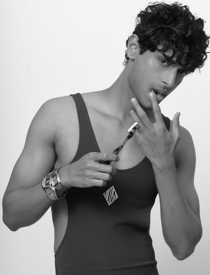 A young adult male in a black tank suit holds a toothbrush near his mouth in a studio setting. Black and white photography.