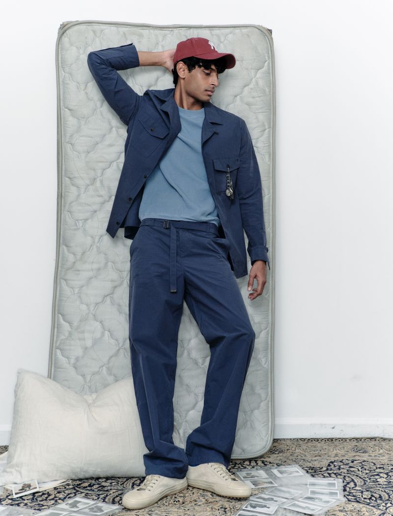 A young adult male lying on a mattress, wearing a navy blue collared jacket, matching pants, and a baseball cap, in a studio setting.