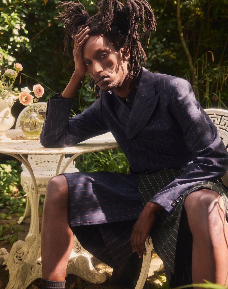 Adult male with dreadlocks sits at white metal table outdoors, wearing a navy pinstripe double-breasted suit in a garden setting.