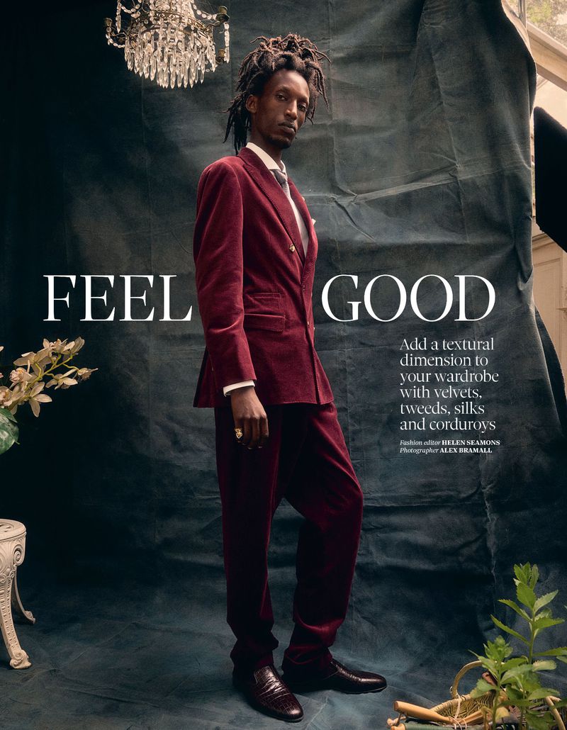 A Black male fashion model poses in a burgundy velvet suit, indoors, next to a vintage lamp and lush plants against a textured backdrop.