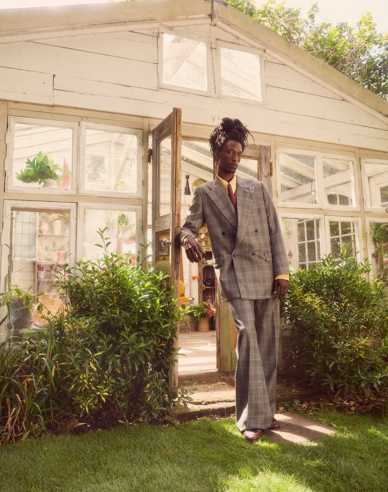 A man in a grey plaid suit leans against a white greenhouse entrance, creating a fashionable and poised outdoor portrait.