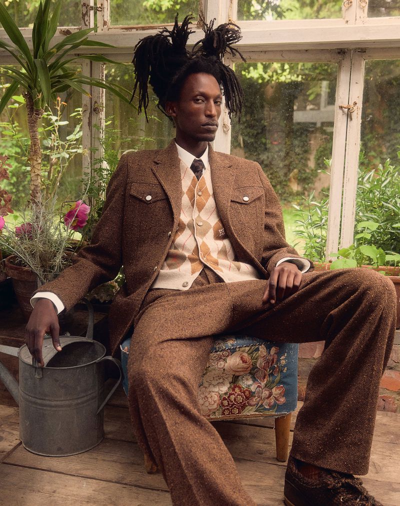 A Black man in a brown suit with an argyle sweater vest sits in a greenhouse on a stool with a floral pattern, next to a watering can.