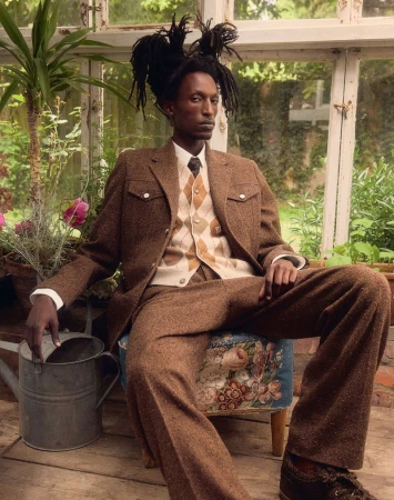 A Black man in a brown suit with an argyle sweater vest sits in a greenhouse on a stool with a floral pattern, next to a watering can.