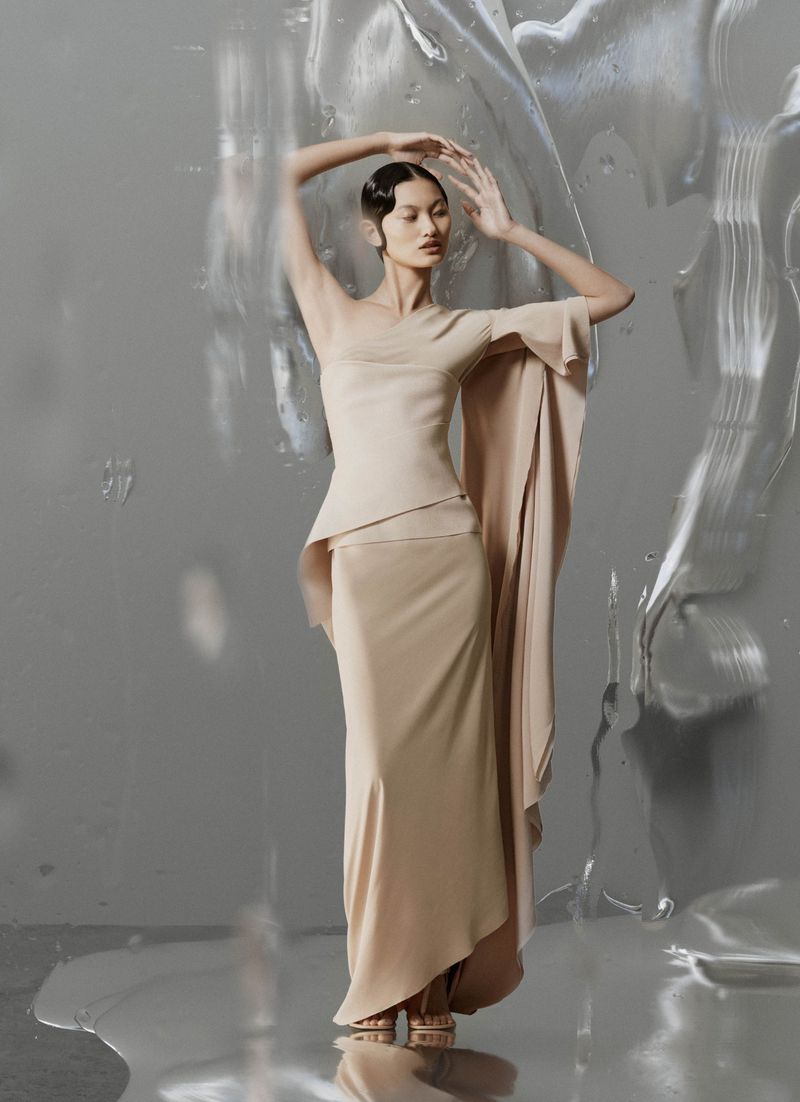 Elegant Asian woman poses in a flowing beige gown against a textured silver backdrop, creating a sophisticated and visually striking image.