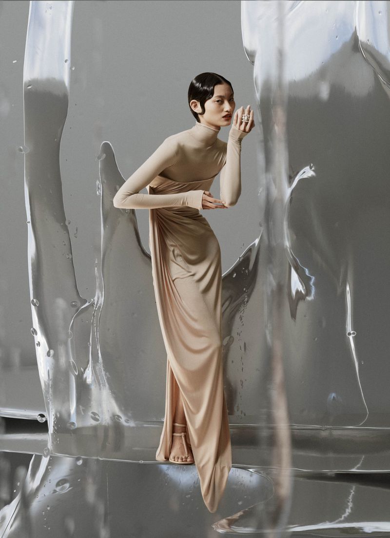 A stylish woman with short hair wears a tan-colored dress, accessorized with rings and open-toed shoes, posing in an indoor photo studio.