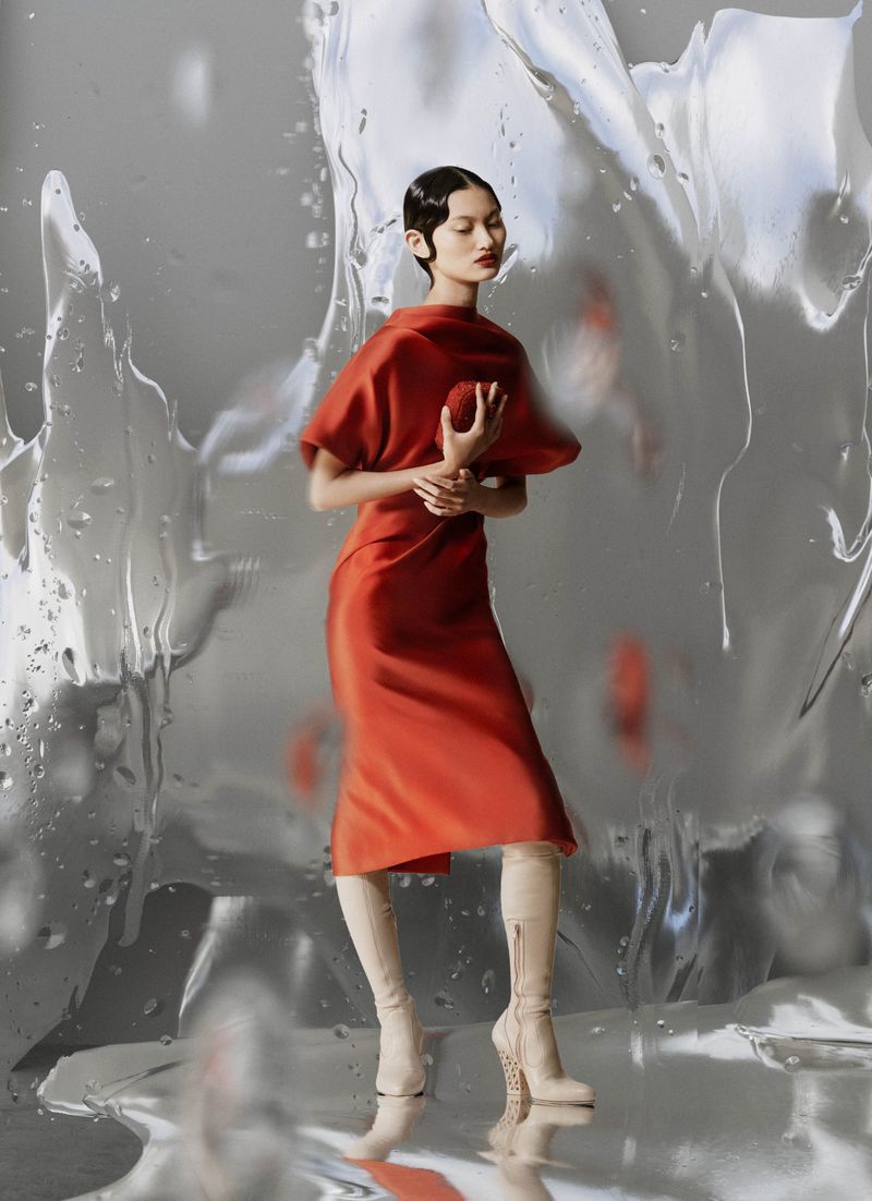Elegant Asian woman in orange dress and beige boots posing against textured silver background in studio fashion photography.