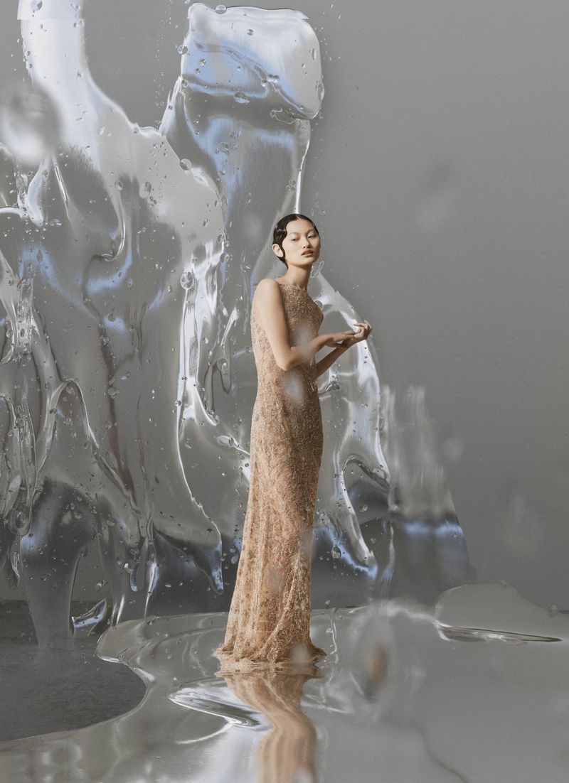 Elegant woman in embellished gown stands near abstract ice sculpture in a fashion photography studio. Focus on the gown and unique set design.