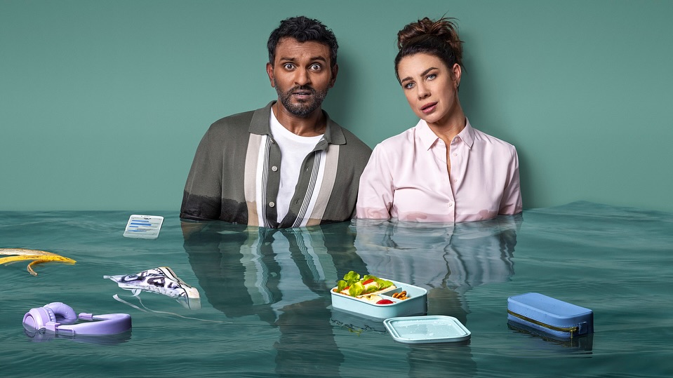 Two adults in waist-deep water surrounded by floating everyday items like a lunchbox, wallet, headphones, and banana peel, against a teal backdrop.