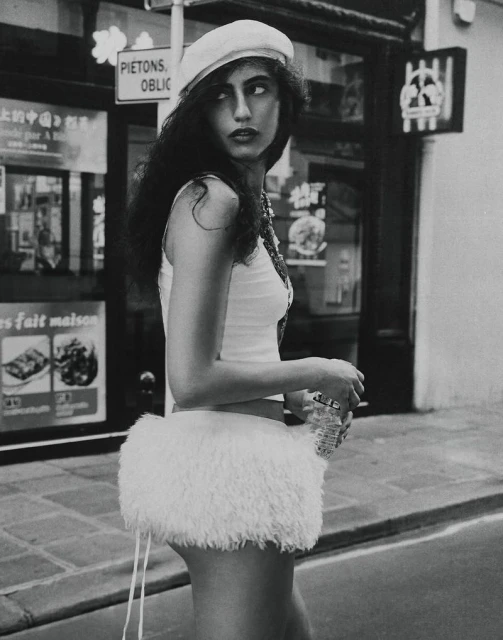 A woman in a white beret and a short white skirt is captured in a monochrome street-style photograph with a storefront backdrop.