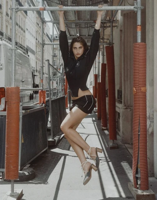 An adult woman hangs from a metal bar in an urban environment, wearing a black outfit and heeled shoes, posing creatively with a neutral expression.