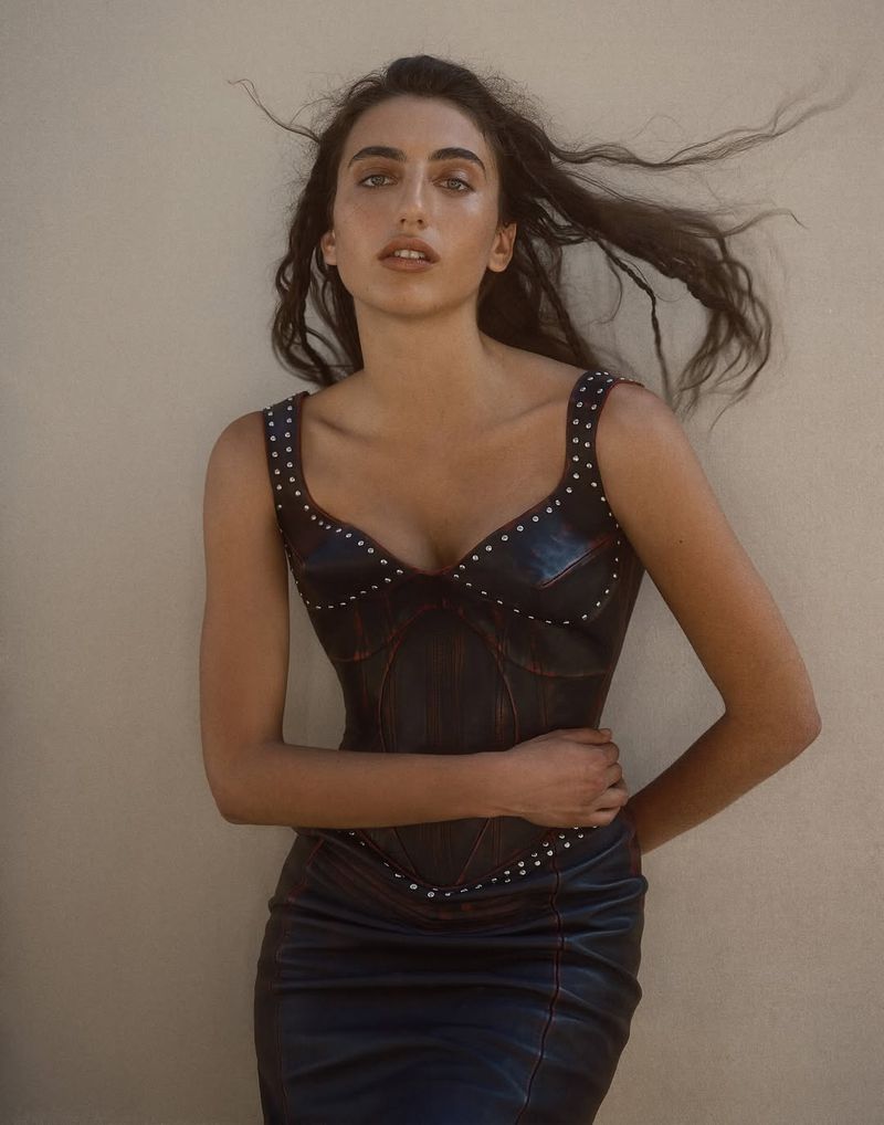 Attractive young woman poses indoors in a leather dress with metal studding. She is fair-skinned with long brown hair and light eye makeup.