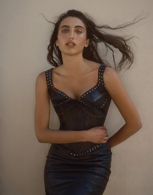 Attractive young woman poses indoors in a leather dress with metal studding. She is fair-skinned with long brown hair and light eye makeup.