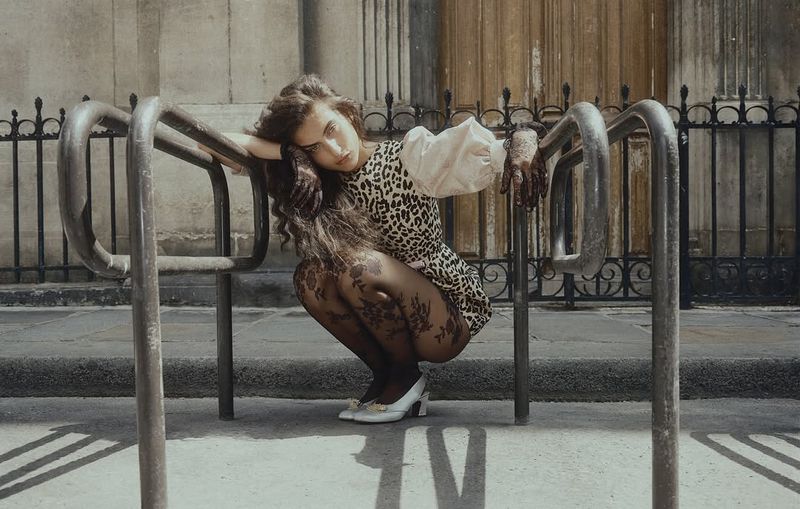 A woman with long curly hair in a leopard-print dress and patterned tights squats between metal barriers in an outdoor urban environment.