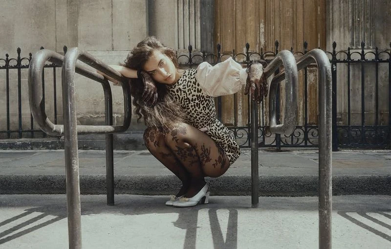 A woman with long curly hair in a leopard-print dress and patterned tights squats between metal barriers in an outdoor urban environment.