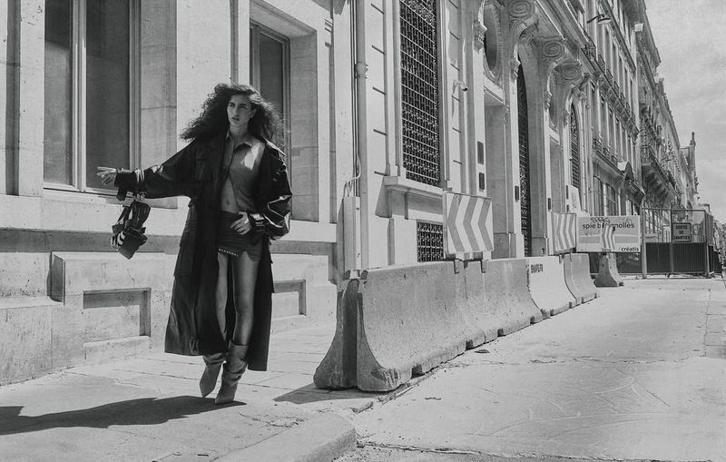 Woman in black coat walks on city sidewalk, carrying bag. Urban fashion shot, architectural details. Monochrome aesthetic.