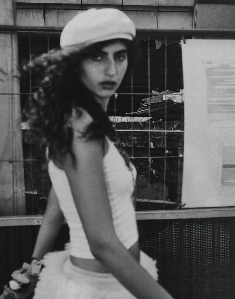 Monochrome portrait of a woman with long curly hair, wearing a beret and cropped top, standing outdoors near a construction fence and posters.