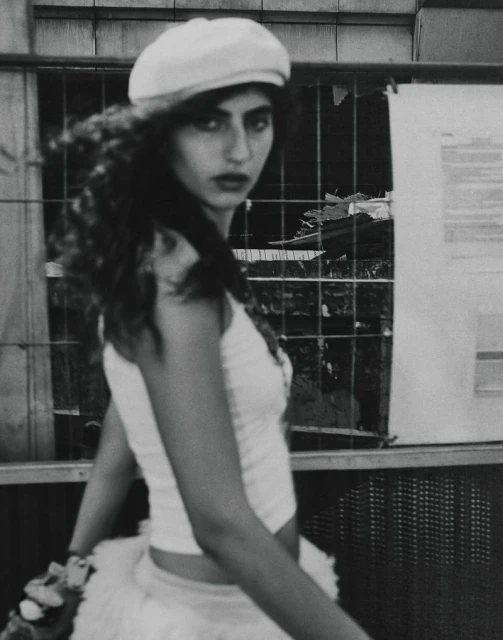 Monochrome portrait of a woman with long curly hair, wearing a beret and cropped top, standing outdoors near a construction fence and posters.